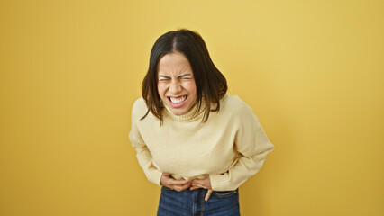 A hispanic woman grimacing with clenched teeth against a yellow background in a casual outfit,...