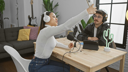 A woman and man taking a selfie in a radio studio with podcasting equipment.