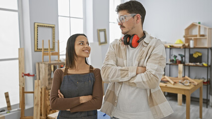 Man and woman smiling in a bright carpentry workshop with woodworking tools and wooden furniture.