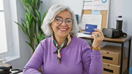 Smiling middle-aged woman holding credit card in a modern office setting, implying financial,...