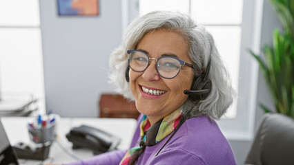 Smiling mature woman with headset working at her desk in a modern office setting, embodying professionalism and friendliness.