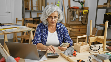 Mature grey-haired woman counting money in a woodworking workshop while using a calculator.