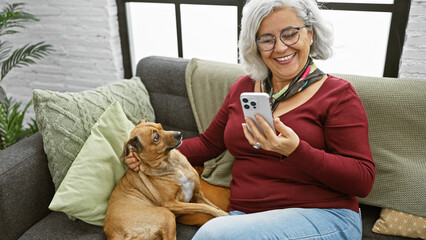 A smiling grey-haired woman enjoys using her smartphone next to her attentive dog on a cozy couch...