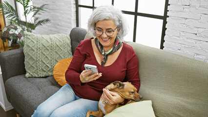 A cheerful mature woman enjoys using her smartphone while cuddling her dog on a cozy sofa indoors.