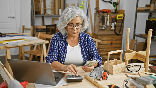 Mature grey-haired woman counting money in a woodworking workshop while using a calculator. - Powered by Adobe
