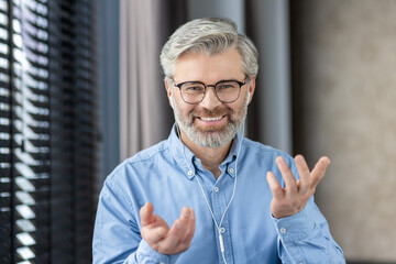 Confident older man with grey beard and glasses, wearing earbuds, engaged in a virtual meeting, smiling and gesturing with his hands.