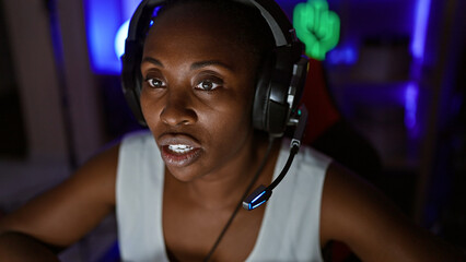 A focused african american woman gaming at night indoors, with vibrant backlighting and wearing a headset.