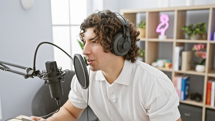 Handsome young hispanic man with curly hair speaking into a microphone in a modern radio studio set
