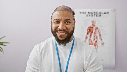 Smiling bearded man with lanyard standing confidently in a clinic with a poster of the muscular...