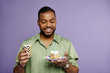 A young, happy African American man with braces holding a plate with birthday cake and a drink on a purple background.