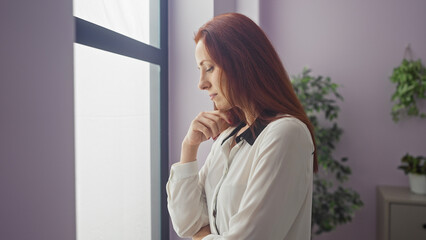 A pensive redhead woman in a white blouse stands contemplatively in a modern apartment room, creating an inviting atmosphere.