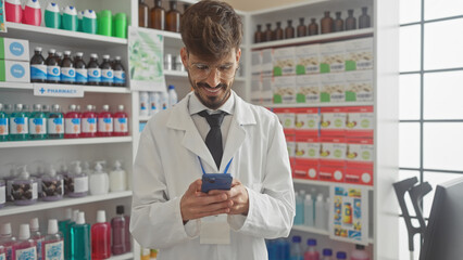 A smiling young man with a beard, wearing glasses and lab coat, using a smartphone in a brightly...