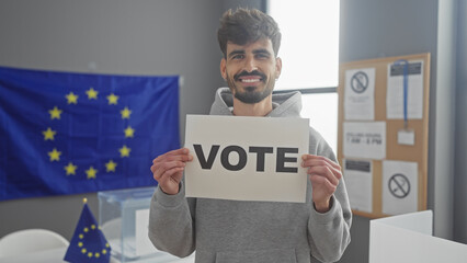 Smiling young man with beard holding a vote sign in a european polling station with eu flags