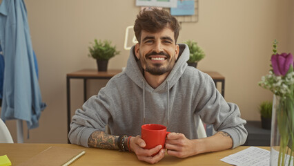 A cheerful young hispanic man with a beard and tattoos holds a red mug, sitting at a table indoors...