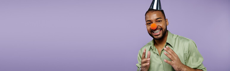 A young African American man with braces on a purple background is happily balancing a clown nose...