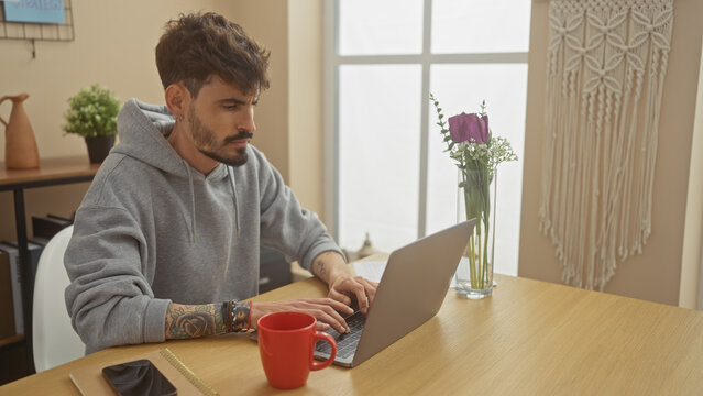 A tattooed young man works on his laptop at a home office table with a red mug nearby in a cozy room. - Powered by Adobe