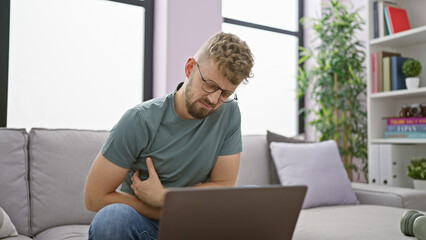 A young bearded man in pain indoors clutching his stomach feeling unwell with a laptop and a water...