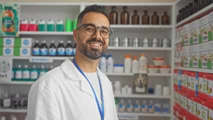 A smiling hispanic male pharmacist stands confidently in a well-stocked, modern pharmacy.