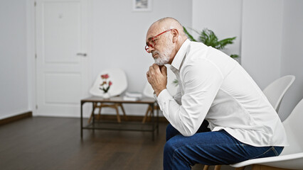 Thoughtful bearded man in white shirt and jeans sitting indoors contemplating in a modern room with...