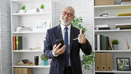 Bald businessman with beard, giving thumbs up in office with smartphone.