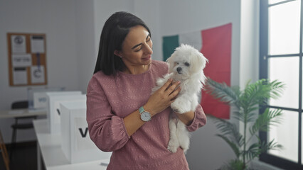 A young woman holds a bichon maltese dog indoors, with the mexican flag in the background of a...