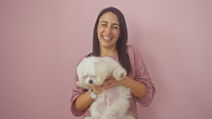 A young hispanic woman laughs joyfully holding a fluffy bichon maltese dog against a pink isolated background.