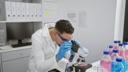 A young man examines specimens under a microscope in a laboratory setting, showcasing research and...