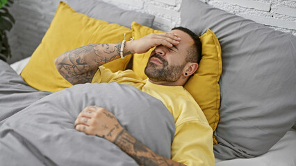 Fototapeta premium Tattooed hispanic man with beard in bedroom feeling headache or stress while lying in bed.