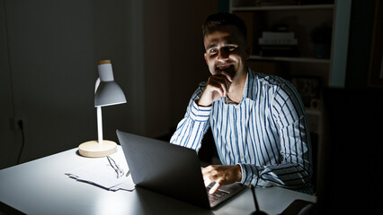 Handsome man working late at night in an office with a laptop and desk lamp, portraying diligence...