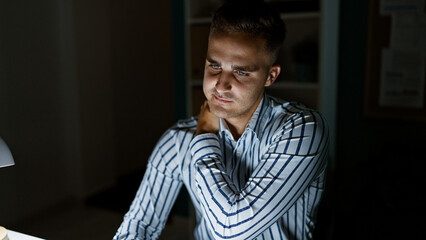 Handsome young man working late in a dark office, illuminated by a desk lamp.
