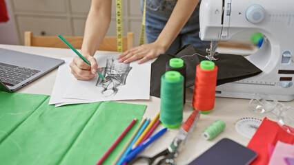 A woman sketches a dress design on paper amid colorful fabrics, threads, and a sewing machine in a tailor's atelier.