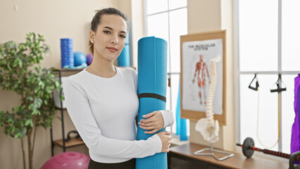 Hispanic woman holding yoga mat in a gym, portraying fitness, exercise, wellness, and health.