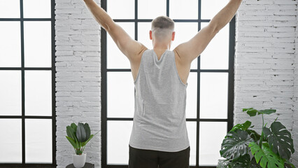 Back view of a young man stretching arms in a bright loft apartment with plants.