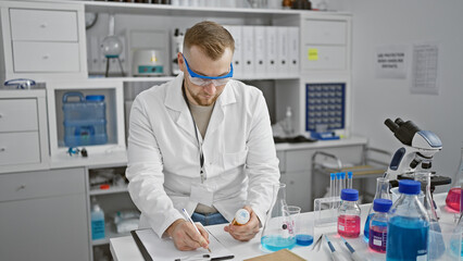 Bearded young man in lab coat and safety glasses examining medicine in laboratory