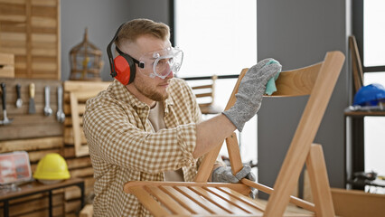 A young, bearded man wearing safety goggles and ear protection meticulously sands a wooden chair in...