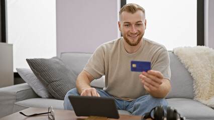 Handsome caucasian man holding credit card using tablet couch home