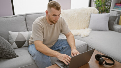 A handsome man using a laptop in a cozy, modern living room setting, depicting work from home.