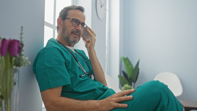 Hispanic male doctor in green scrubs listening to a voice message in a bright hospital room