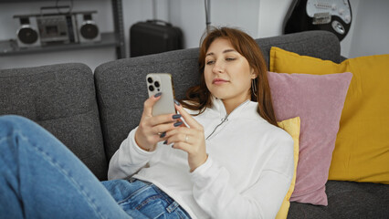 A relaxed young woman lounges on a couch using her smartphone in a cozy living room setting.