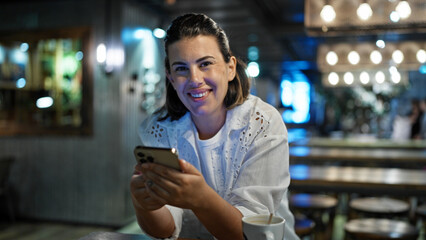 Beautiful young hispanic woman using smartphone smiling at cafeteria