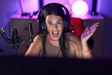 Young hispanic woman playing video games wearing headphones celebrating achievement with happy...
