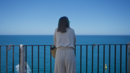 A young hispanic woman stands from behind at a scenic viewpoint in polignano a mare, puglia, italy, gazing at the calm blue sea under a clear sky.