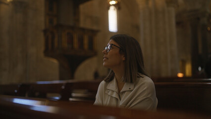 A young hispanic woman sits in contemplation inside a historic church in italy, bathed in the soft light streaming through a tall stained glass window.