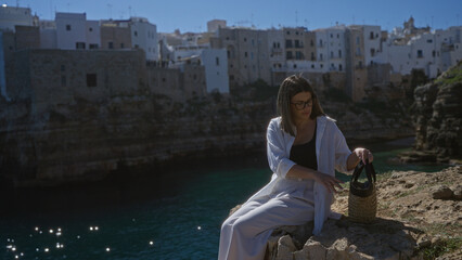 Hispanic woman seated on a rock cliff overlooking the sea in polignano a mare, puglia, italy, under a clear blue sky with picturesque whitewashed buildings in the background.