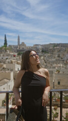 Fototapeta premium A beautiful young hispanic woman enjoying a sunny day at a viewpoint in the old town of matera, basilicata, italy, with historic buildings and a clear blue sky as the backdrop.