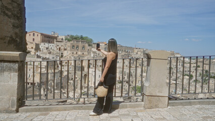 A young, beautiful hispanic woman takes a selfie in the old town of matera, basilicata, italy, capturing the historic cityscape from a scenic viewpoint under a clear, blue sky.