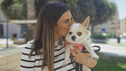 A beautiful young hispanic woman holding and kissing her pet chihuahua in an urban park on a sunny day.