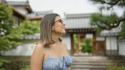 Hispanic female, radiating beauty and joy with glasses, looking around with cheerful expression at kyoto temple, standing confidently amidst japanese architecture, basking in the serenity.