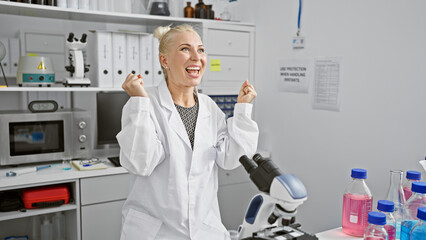 Attractive young blonde woman scientist celebrating a winning discovery, microscope analysis in a bustling lab brings a joyful smile!