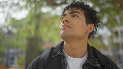 Young hispanic man with curly hair looking upward in a contemplative manner at an urban park setting.